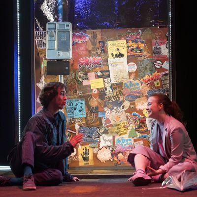 Two actors sit on the floor of the set in front of a phone booth covered in posters and stickers